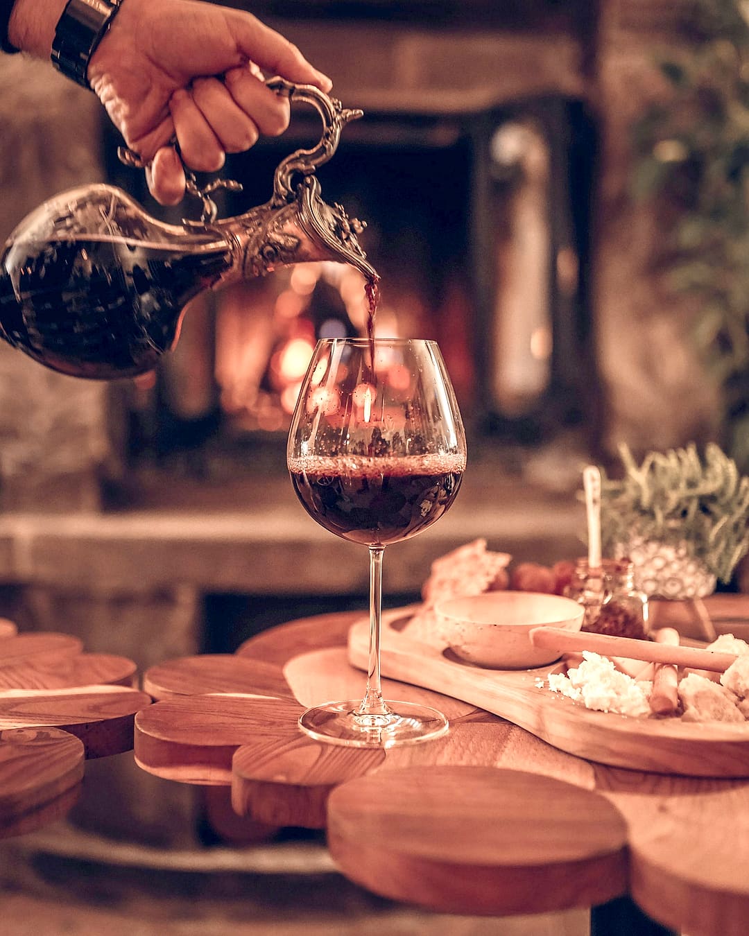 Crno vino toči se iz vrča u čašu, iza je kamin.
